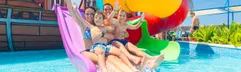 Een lachende familie met kinderen glijdt samen van de brede waterglijbaan in het zwembad van Royal Son Bou op Menorca.