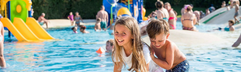 Twee blije kinderen spelen samen in het ondiepe water van het kinderbad op Camping Union Lido, met kleurrijke speeltoestellen op de achtergrond.