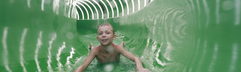 Een jongen glijdt met een grote glimlach door een groene, verlichte waterglijbaantunnel in de Aqua Mundo van Center Parcs Zandvoort.