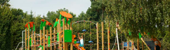 Grote buitenspeeltuin bij De Wije Werelt met houten klimtoestellen, een glijbaan en een grote zandbak, omringd door bomen.