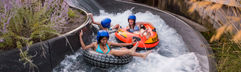 Drie kinderen met helm op zitten samen in een grote opblaasbare band en glijden door de wildwaterbaan, één van de spectaculaire glijbanen van Landal Hof van Saksen.