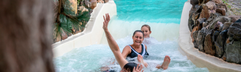 Vrolijk zwaaiend gezin laat zich meevoeren door de stroomversnelling van de wildwaterbaan in het subtropische zwembad van Landal Vennenbos.