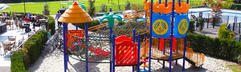 Overzicht van de omheinde en ruime buitenspeeltuin met een groot, kleurrijk speeltoestel en veel zand om in te spelen.