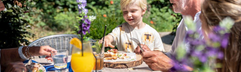 Een lachende familie met kinderen geniet van een lunch buiten aan een houten tafel in de tuin van hun vakantiehuis.