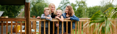Een lachend gezin met drie kinderen leunt over de houten balustrade van de veranda van hun safaritent op Vakantiepark Sallandshoeve.