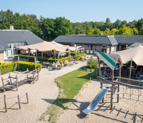 Zonnig terras met uitzicht op de speeltuin met zand en een glijbaan bij de centrumvoorzieningen van kindvriendelijk vakantiepark Bospark Lunsbergen.