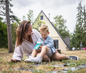 Een moeder en haar zoontje zitten op een kleed in het gras voor hun bungalow te spelen met een bellenblaas op vakantiepark De Katjeskelder.