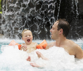 Een lachende vader en zoon genieten samen in het bruisende bubbelbad van de Aqua Mundo bij Het Meerdal, met op de achtergrond een grote waterval.