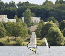 Een windsurfer op het water van het Bostalmeer, met op de achtergrond de vakantiehuizen van Center Parcs Park Bostalsee.