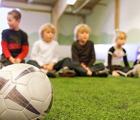 Kinderen luisteren naar de coach tijdens een voetbalclinic in de sporthal van Center Parcs Park Hochsauerland, met een voetbal op de voorgrond.