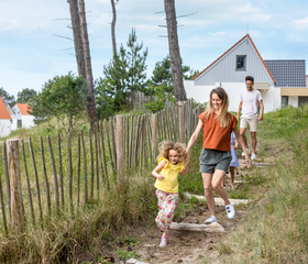 Een gezin wandelt hand in hand over een pad door het duinlandschap op vakantiepark Center Parcs Zandvoort, met een cottage op de achtergrond.