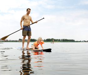 Een vader staat met een peddel op een sup-board terwijl zijn dochter voorop zit, op het grote Eemmeer bij Center Parcs De Eemhof.