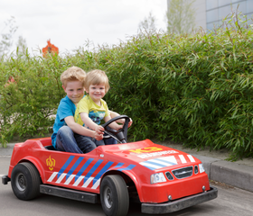 Twee blije jongens rijden samen in een rode speelgoed-brandweerauto door het Kids Verkeerspark bij Center Parcs De Huttenheugte.