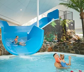 Een peuter glijdt van een brede blauwe glijbaan het kinderbad in van het subtropische zwembad op Recreatiepark De Leistert, waar een vader hem opwacht.
