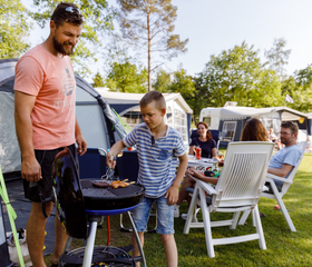 Een gezellig gezin dat aan het barbecueën is bij hun kampeerplek op RCN de Roggeberg, waarbij de zoon de vader helpt bij de BBQ.
