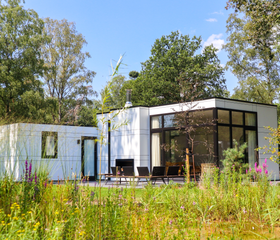Een moderne, witte design vakantiewoning met een terras, omgeven door wilde bloemen en het groen van de Veluwe op Droompark Hooge Veluwe.