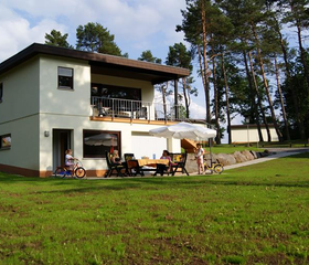 De buitenkant van een moderne vakantiewoning in Feriendorf Felsenhof, met een ruim grasveld en een terras waar gasten zitten.