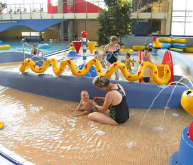 Een baby speelt met moeder in het ondiepe kinderbad van het Brockenbad, met een gele waterslang en andere speelelementen.
