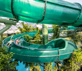 Een bezoeker glijdt met hoge snelheid door een lange, groene en deels doorzichtige waterglijbaan die tussen de tropische planten van Aqua Mundo slingert.