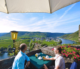 Twee volwassenen genieten van een drankje op het terras van Hotel Burgfrieden met een spectaculair panoramisch uitzicht over de kronkelende Moezelrivier en groene wijngaarden.