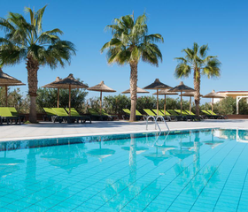 Rustig zwembad omringd door palmbomen en comfortabele ligbedjes met parasols, ideaal voor ontspanning in de Griekse zon.