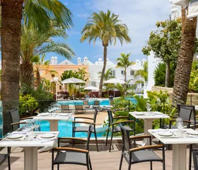 Gasten genieten van hun maaltijd aan tafels op het buitenterras van het restaurant, onder een pergola en met uitzicht op de tropische tuin.