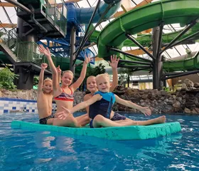Vier vrolijk zwaaiende kinderen hebben plezier op een grote, drijvende mat in het zwembad van het subtropisch zwemparadijs op het kindvriendelijke vakantiepark Landal De Lommerbergen.