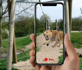 Een smartphone toont een sabeltandtijger via een augmented reality app, waardoor het lijkt alsof het prehistorische dier op het vakantiepark rondloopt.