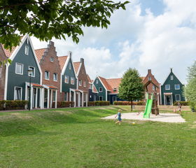 Een speeltuintje met glijbaan op een groot grasveld, omringd door karakteristieke vakantiehuizen in Volendamse stijl