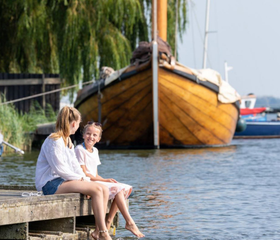 Een gezin zit met de voeten boven het water op een steiger in de haven, met een klassiek houten schip op de achtergrond.