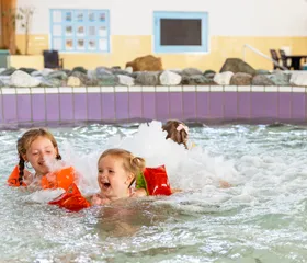 Twee lachende meisjes met zwembandjes spelen en spetteren in het ondiepe, bruisende kinderbad van het zwembad op Vakantiepark Port Greve.