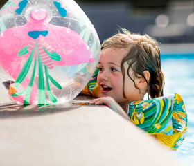 Lachend meisje met zwembandjes leunt op de rand van het zwembad en houdt een vrolijke strandbal vast op Recreatiepark Beekbergen.
