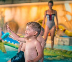 Lachende jongen spettert vrolijk in het binnenzwembad van Resort Kaatsheuvel, omringd door speelelementen.
