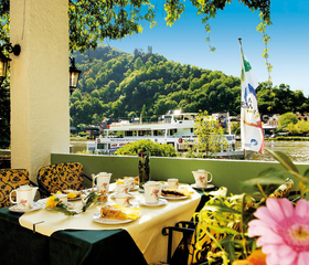 Het terras van Hotel Bellevue met een gedekte tafel en uitzicht op de kasteelruïne Grevenburg en de wijngaarden van de Moezel.