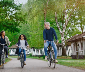 Een gezin van drie fietst over een pad op Vakantiepark Beekse Bergen, langs de accommodaties met zebraprint.