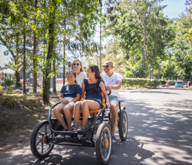 Een gezin van vier personen rijdt vrolijk op een familiefiets (skelter) door de bosrijke omgeving van Vakantiepark Bospark 't Wolfsven.