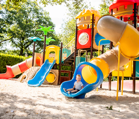 Een kindje glijdt van een kleurrijk speeltoestel in de buitenspeeltuin van Vakantiepark Leuvert, omringd door bomen.