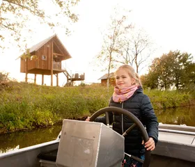 Een meisje staat aan het roer van een fluisterbootje en vaart over de rivier de Regge bij Vakantiepark Molke, met een boomhut op de achtergrond.