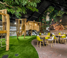 Het indoor speelparadijs van Vakantiepark Schaijk, met houten klimtoestellen en een gezellig terras voor ouders.