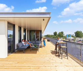 Een gezin ontspant op het zonnige en ruime houten terras van een moderne waterlodge direct aan het water in Zeewolde.