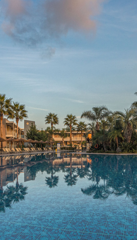 Het grote zwembad van Blue Lagoon Resort op Kos, met palmbomen die prachtig weerspiegelen in het kalme water tijdens zonsondergang.