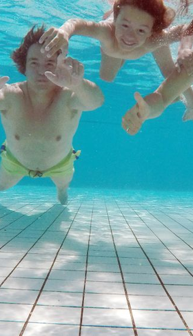 Lachende kinderen zwemmen en zwaaien onder water in het heldere zwembad van Bungalowpark Hoenderloo op de Veluwe.