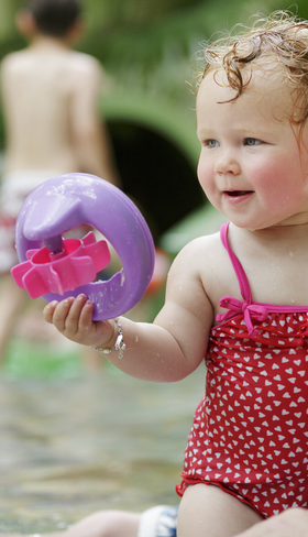 Een lachende dreumes in een rood badpakje speelt met een bal in het ondiepe water van het subtropische zwemparadijs Aqua Mundo bij Center Parcs Bispinger Heide.