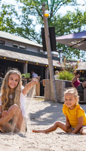 Twee kinderen spelen vrolijk in het witte zand op het strandterras van het restaurant bij Landal Klein Oisterwijk in Noord-Brabant.