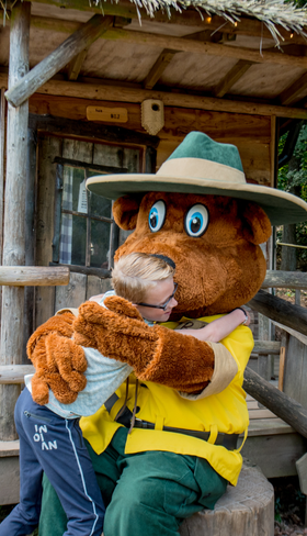 Een kind geeft een dikke knuffel aan mascotte Bollo de Beer, die bij een houten speelhuisje zit op vakantiepark Landal Rabbit Hill op de Veluwe.