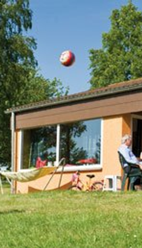 Een meisje gooit een bal in de lucht op het grasveld voor de bungalow op Landal Warsberg, terwijl haar familie op het terras zit.