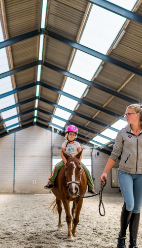 Een meisje met een cap op krijgt paardrijles op een pony in de overdekte manege van Landgoed de Scheleberg op de Veluwe.