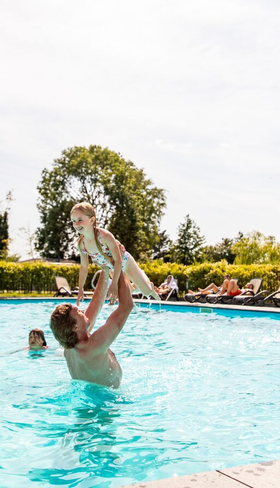 Een vader gooit zijn kind lachend in de lucht in het verwarmde buitenzwembad van Parc de IJsselhoeve, vol waterpret.