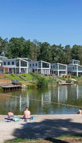 Gasten zwemmen en zonnen bij de recreatieplas met zandstrand op Resort Brunssummerheide in Limburg, met uitzicht op de luxe waterwoningen.