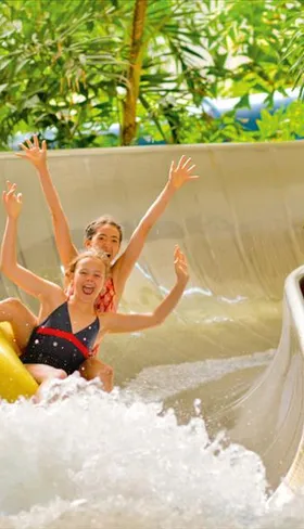 Twee blije meiden roetsjen met hun armen in de lucht in een gele band van een brede waterglijbaan in het zwemparadijs van Tropical Islands.